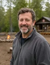 A camp host with a grey beard in a fleece jacket standing in front of a camp fire and a timber hut