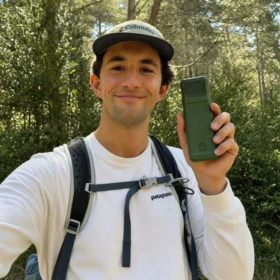 A bearded man in a cap and white jumper with a hiking pack, standing in sunlit forest and holding up a MoskiCell unit