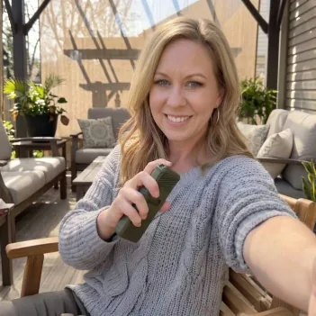 A woman in a grey knitted cardigan holding a MoskiCell unit on a covered patio with outdoor furniture and pot plants behind her