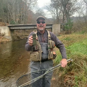 An angler in waders and a fishing vest standing in a river holding a landing net and a MoskiCell unit