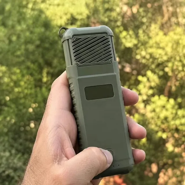 A hand holding the switched on MoskiCell unit in front of sunlit green foliage with a thumb resting on the power button